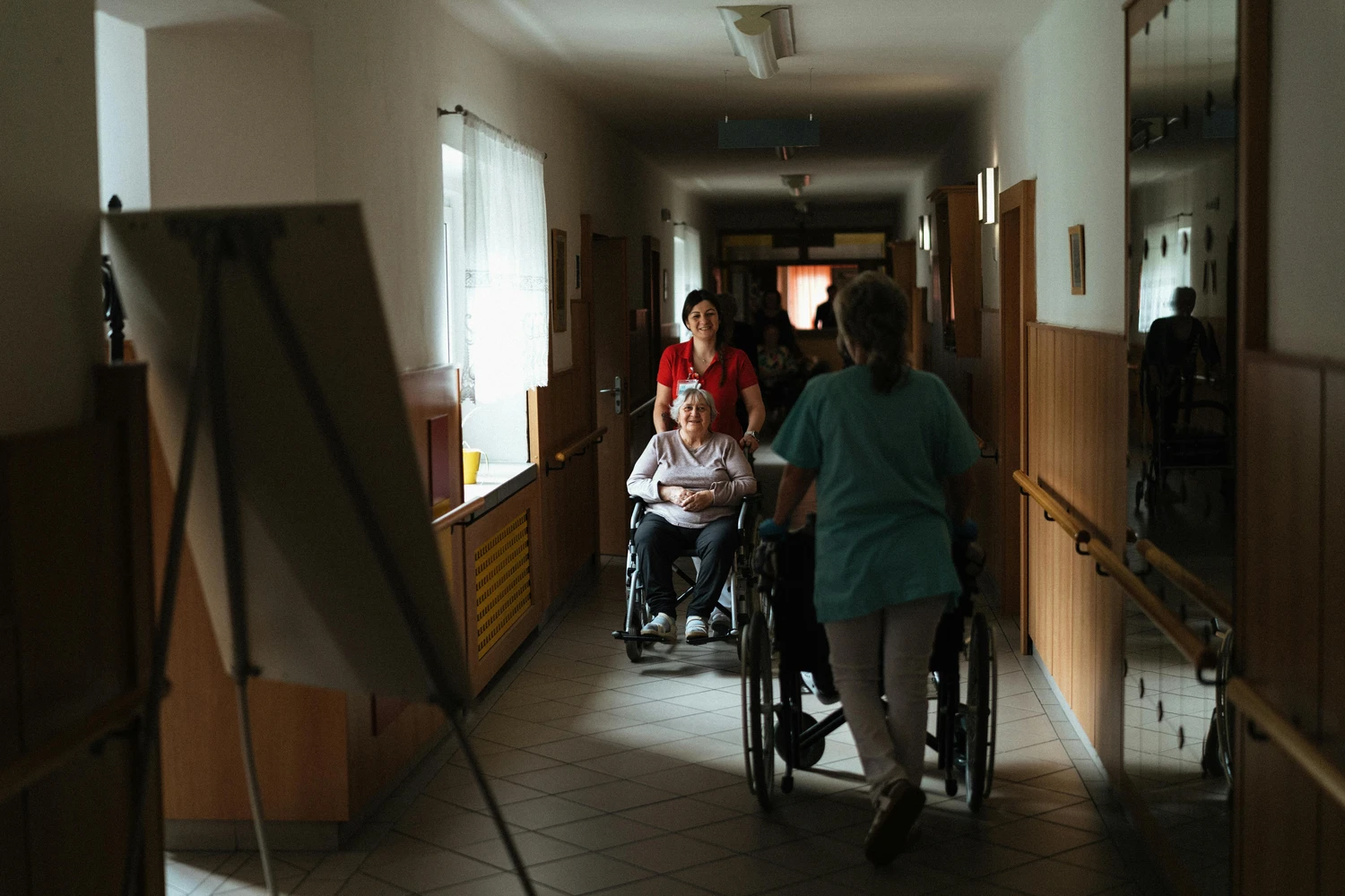 AB Ambulanter Pflegedienst - Traunstein Ambulanter Betreuungs und Pflegedienst - Eine Pflegekraft hilft einem älteren Bewohner im Rollstuhl auf dem Flur einer Langzeitpflegeeinrichtung.